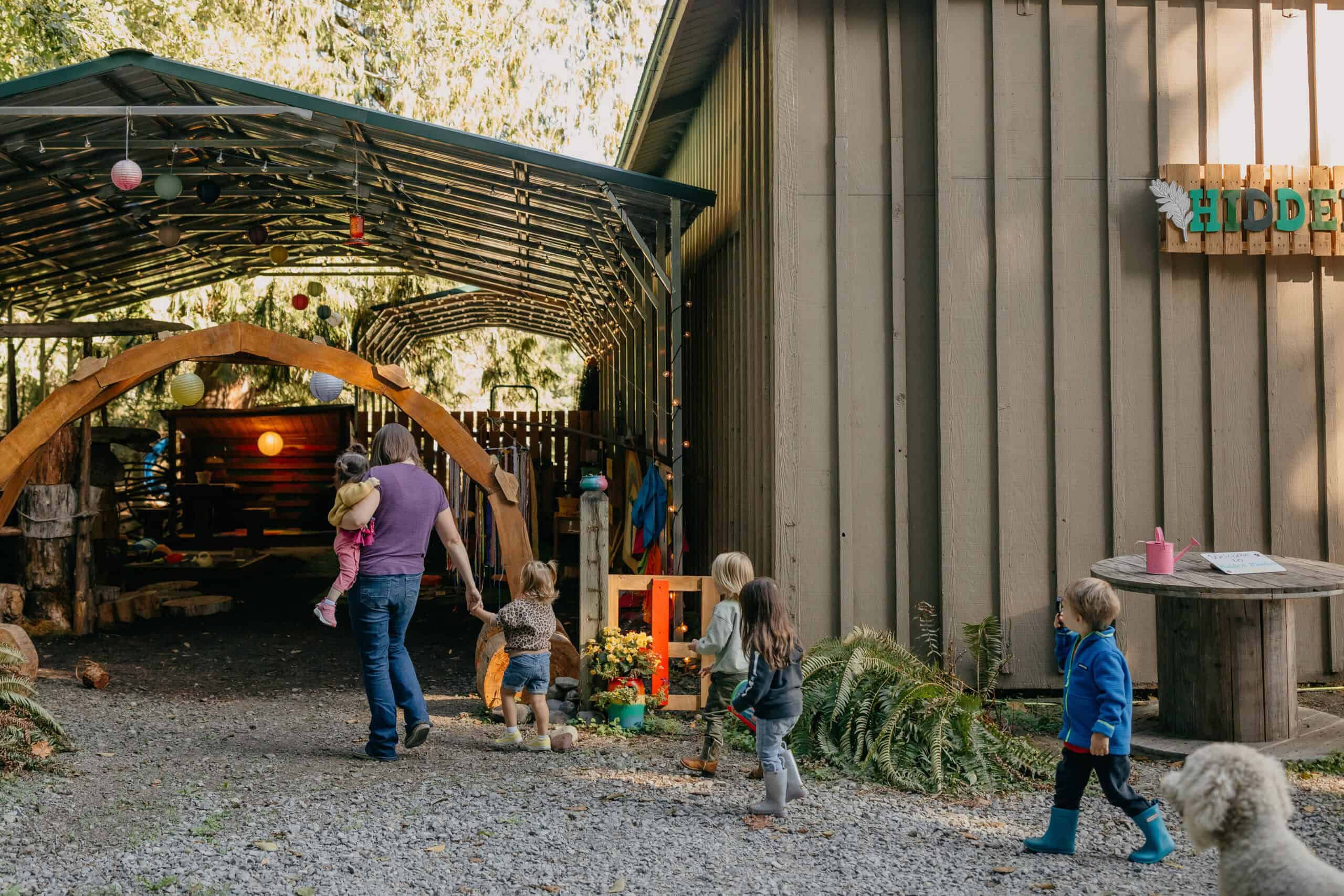 Molly walking into an outdoor space at Hidden Meadow School carrying a child with children in line behind her.