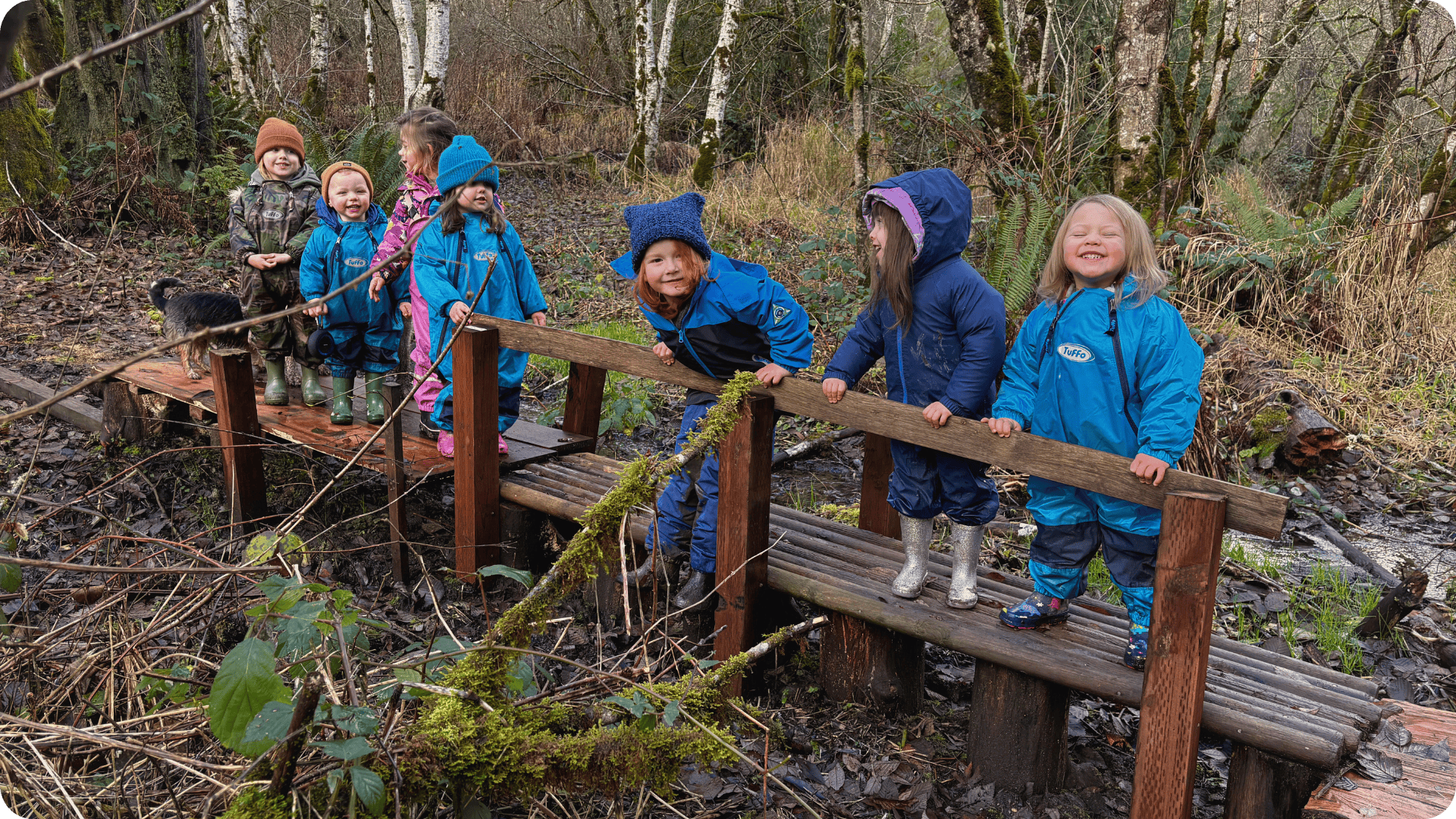 Children standing on bridge over creek in a line smiling at Hidden Meadow School