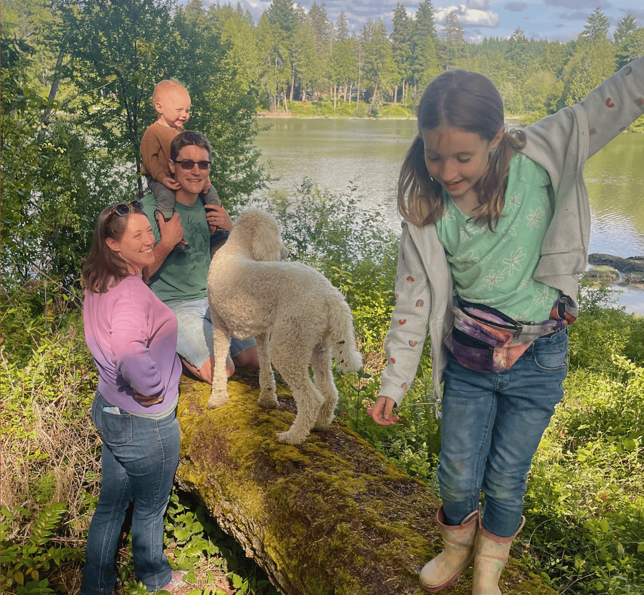 Molly O'rear and family. Molly smiling with daughter balancing on log with Jake holding Quincy on his shoulders and their dog Wilma also on the log. The background is the forest of Hidden Meadow School with the water inlet behind them.