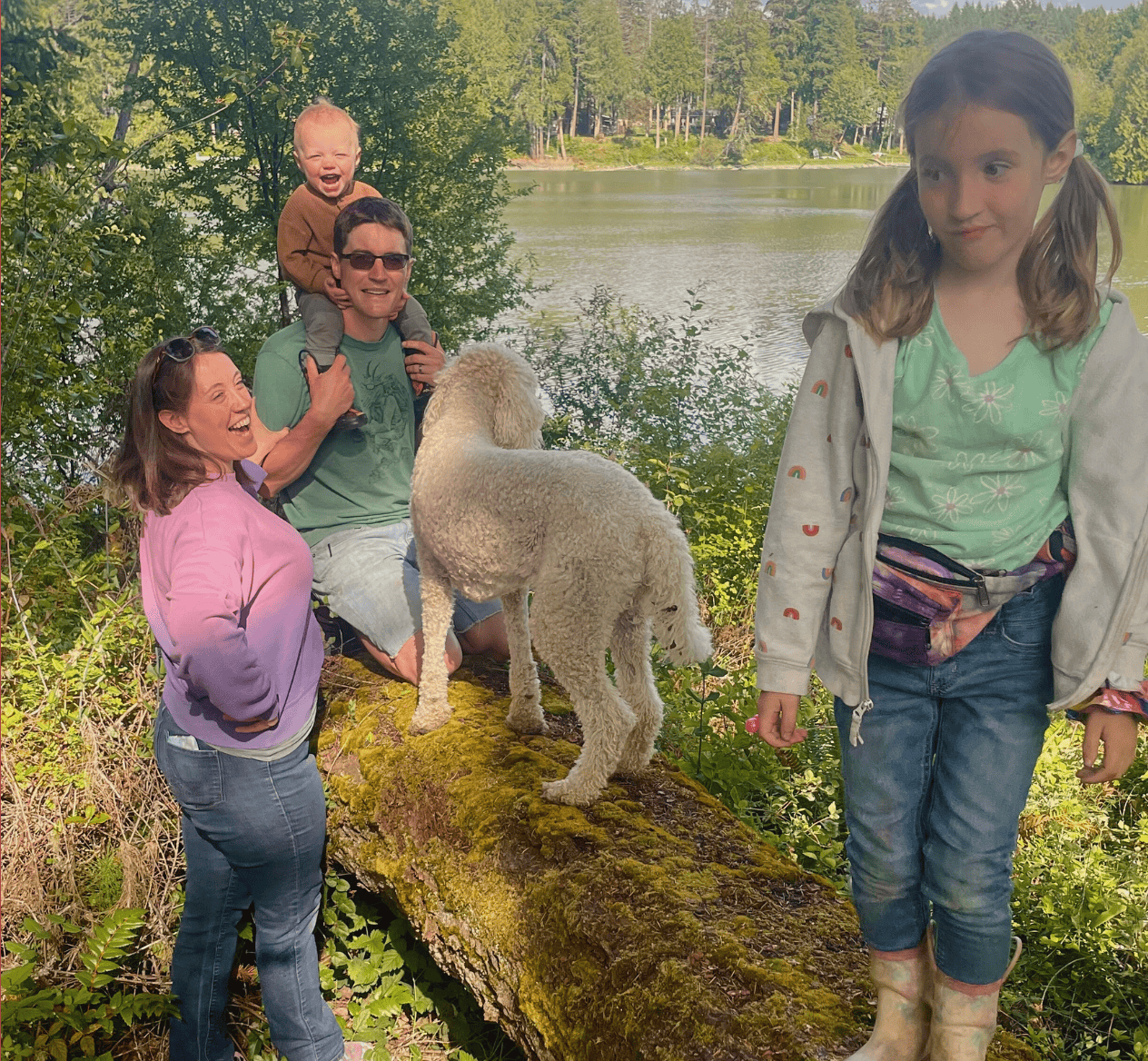Molly O'rear and family. Molly laughing with daughter balancing on log with Jake holding Quincy on his shoulders and their dog Wilma also on the log. The background is the forest of Hidden Meadow School with the water inlet behind them.