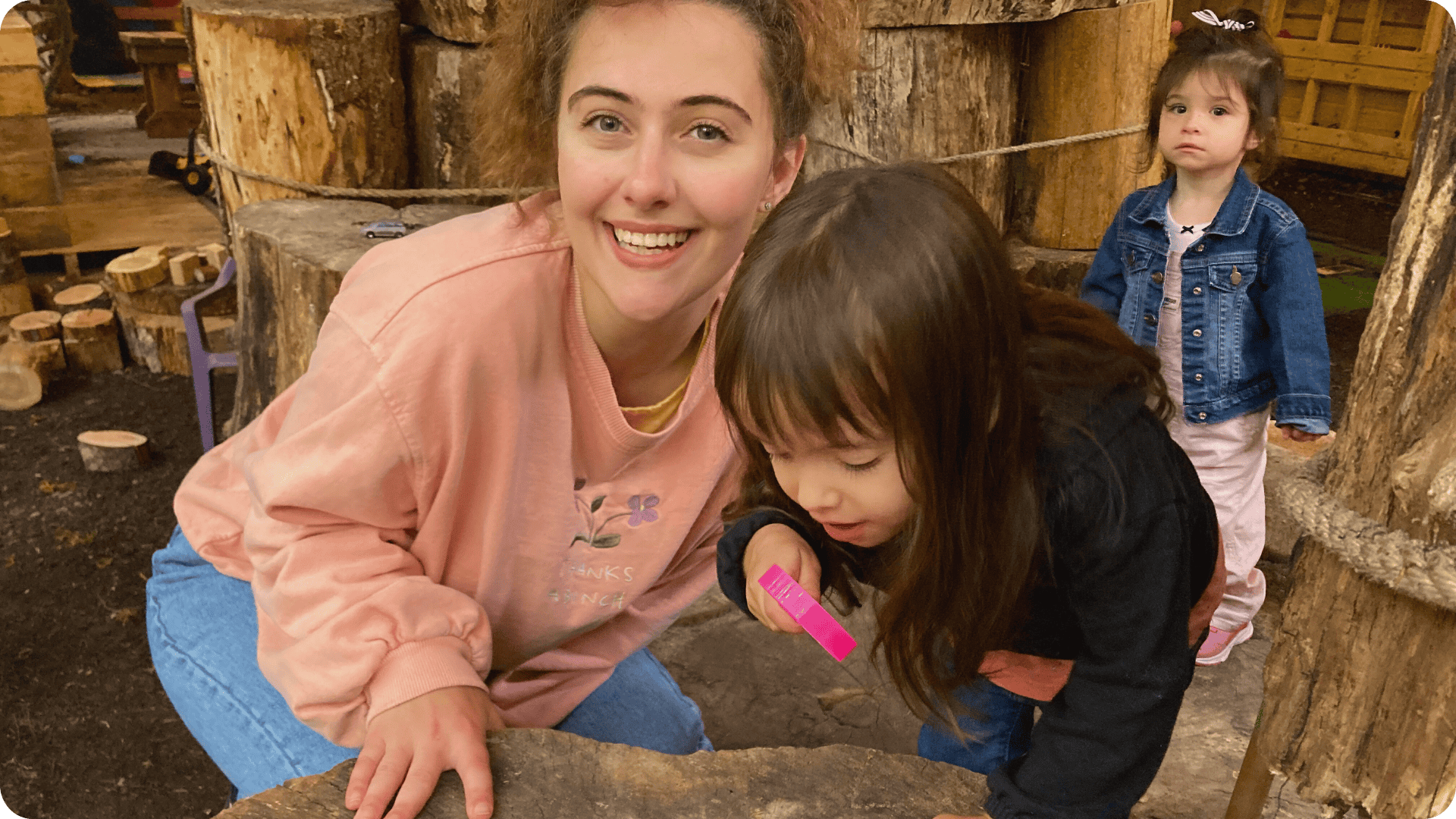 Dani smiling at the camera while a child investigates something on a log with a magnifying glass